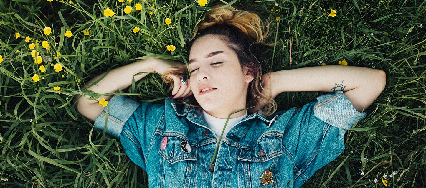 rapariga deitada de barriga para cima sobre a erva no campo, com casaco de ganga, e com os braços à altura da cara