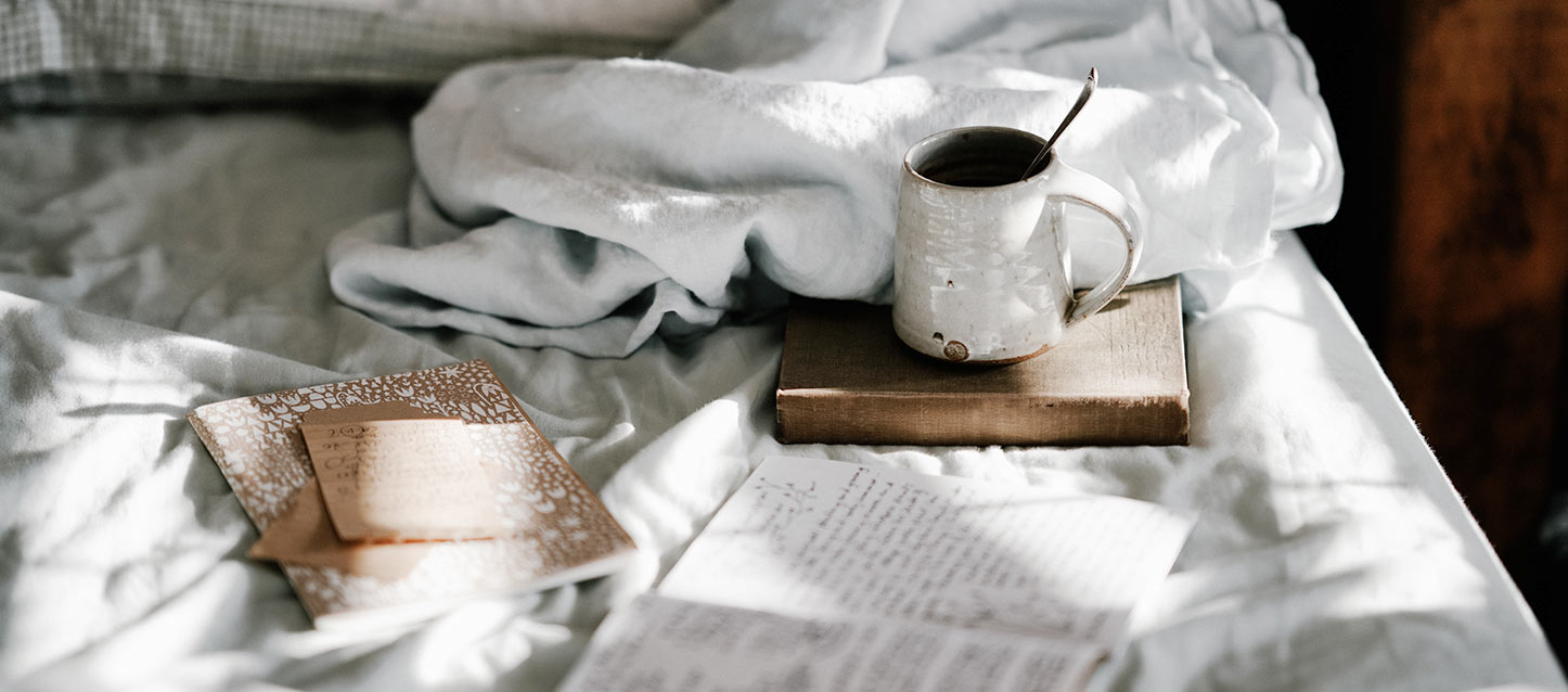 cama com taça de café sobre um livro, bloco para notas, e outro bloco aberto com apontamentos