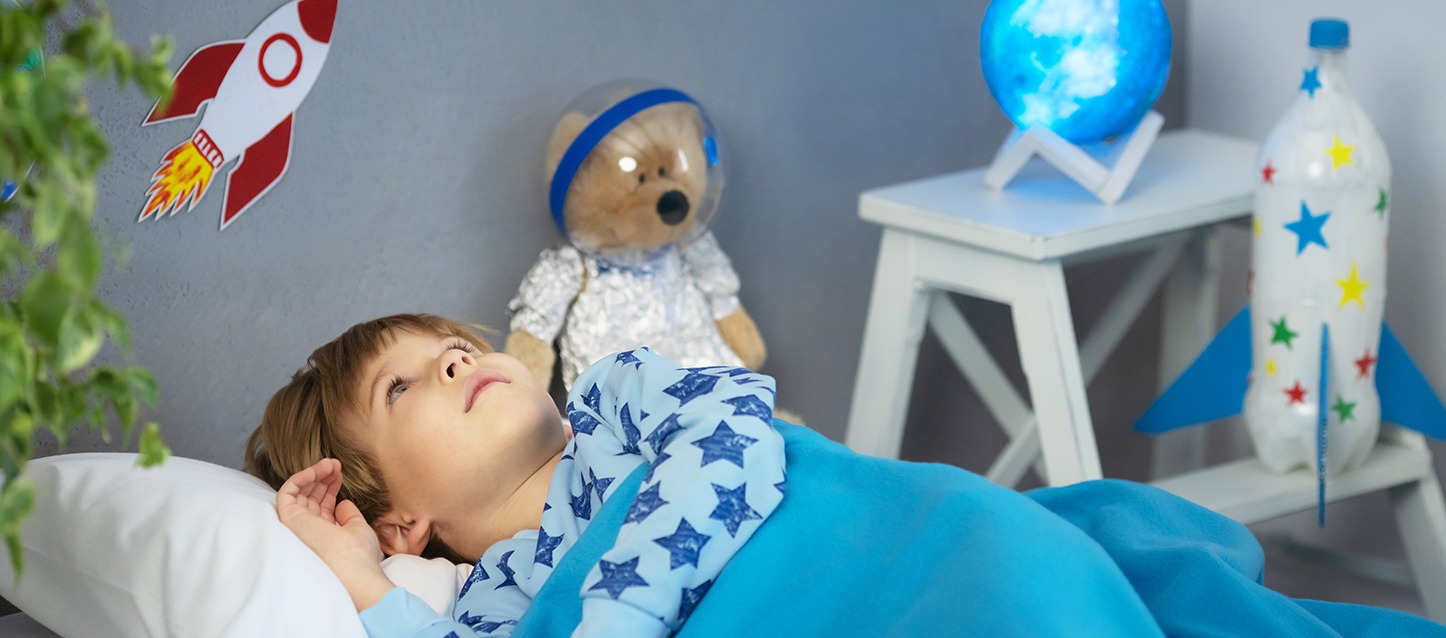menino pequeno de cabelo castanho, com pijama azul claro e estrelas azuis-escuras, metido na cama e tapado com uma manta azul, a olhar para o teto