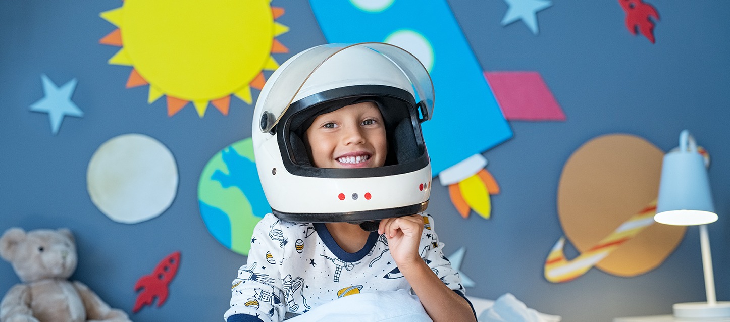 menino pequeno com capacete de moto de cor branca e grande, com pijama branco com motivos espaciais, e de fundo uma parede azul com decorações espaciais