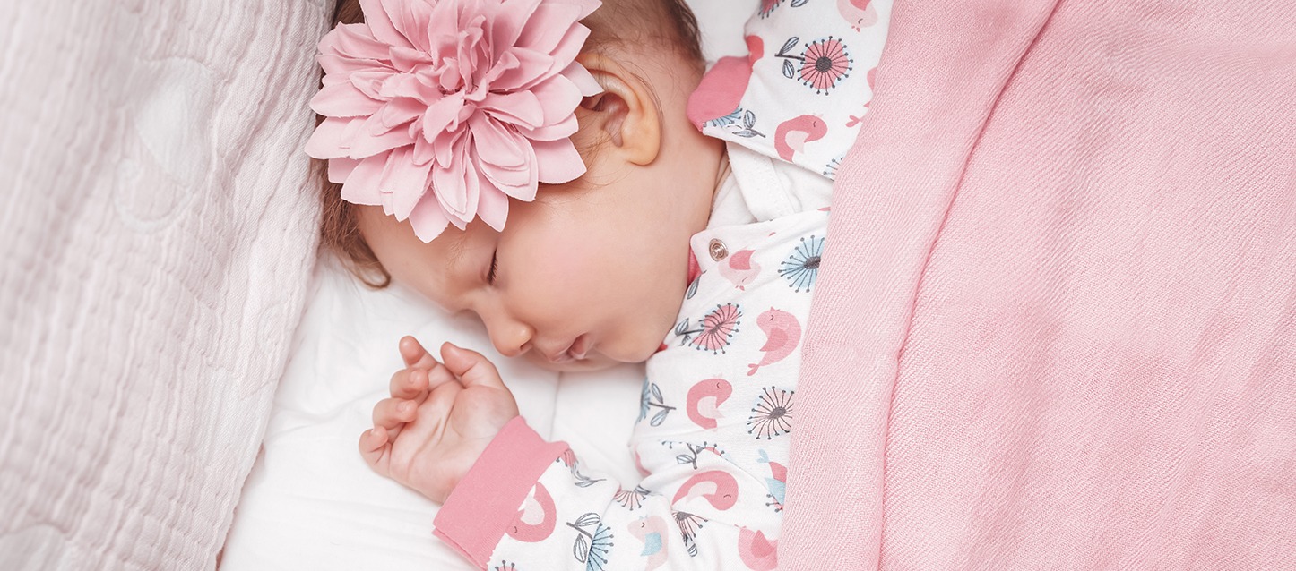 menina bebé a dormir de barriga para cima, com umas flores decorativas na cabeça, e tapada com uma manta rosa