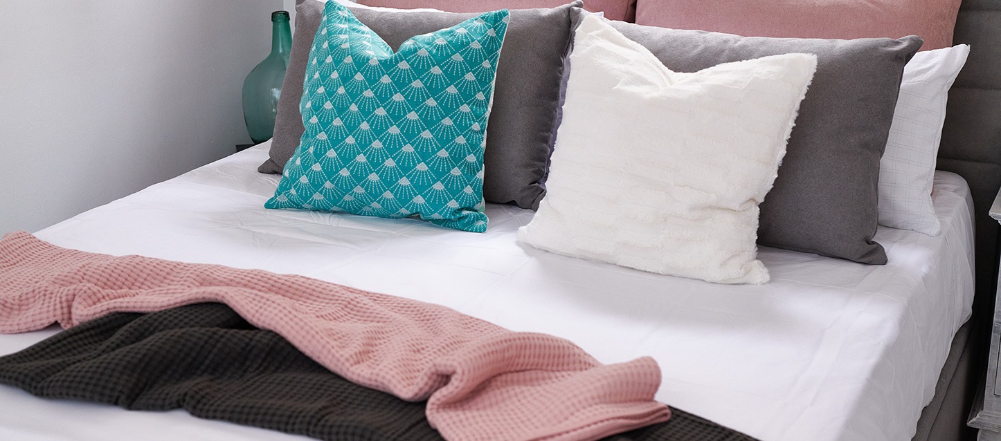 cama com duas mantas dobradas em cima; uma rosa e outra castanha, 6 travesseiros e 2 almofadas