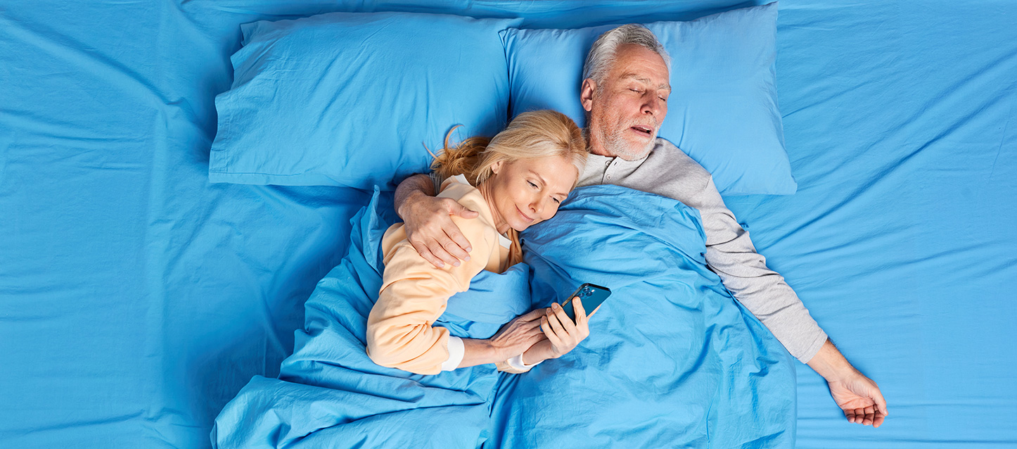 casal de pessoas idosas na cama, em que ele está a dormir e ela está apoiada no seu ombro a olhar para o telemóvel