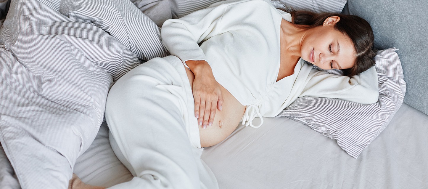 mulher de cabelo moreno, grávida, deitada de lado na cama a tocar e a olhar a sua barriga