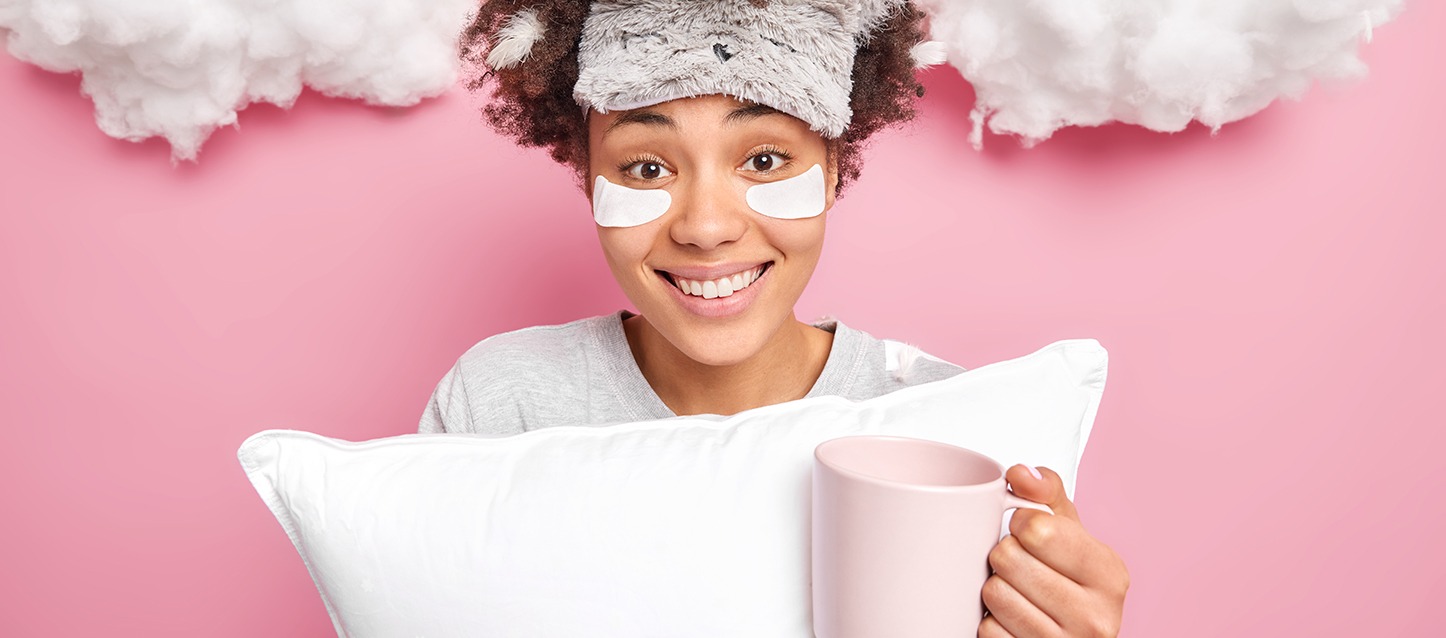 rapariga morena de cabelo afro com adesivos anti-olheiras brancos, máscara para os olhos na testa, pijama cinza, abraçando uma almofada e segurando uma taça de café de cor rosa