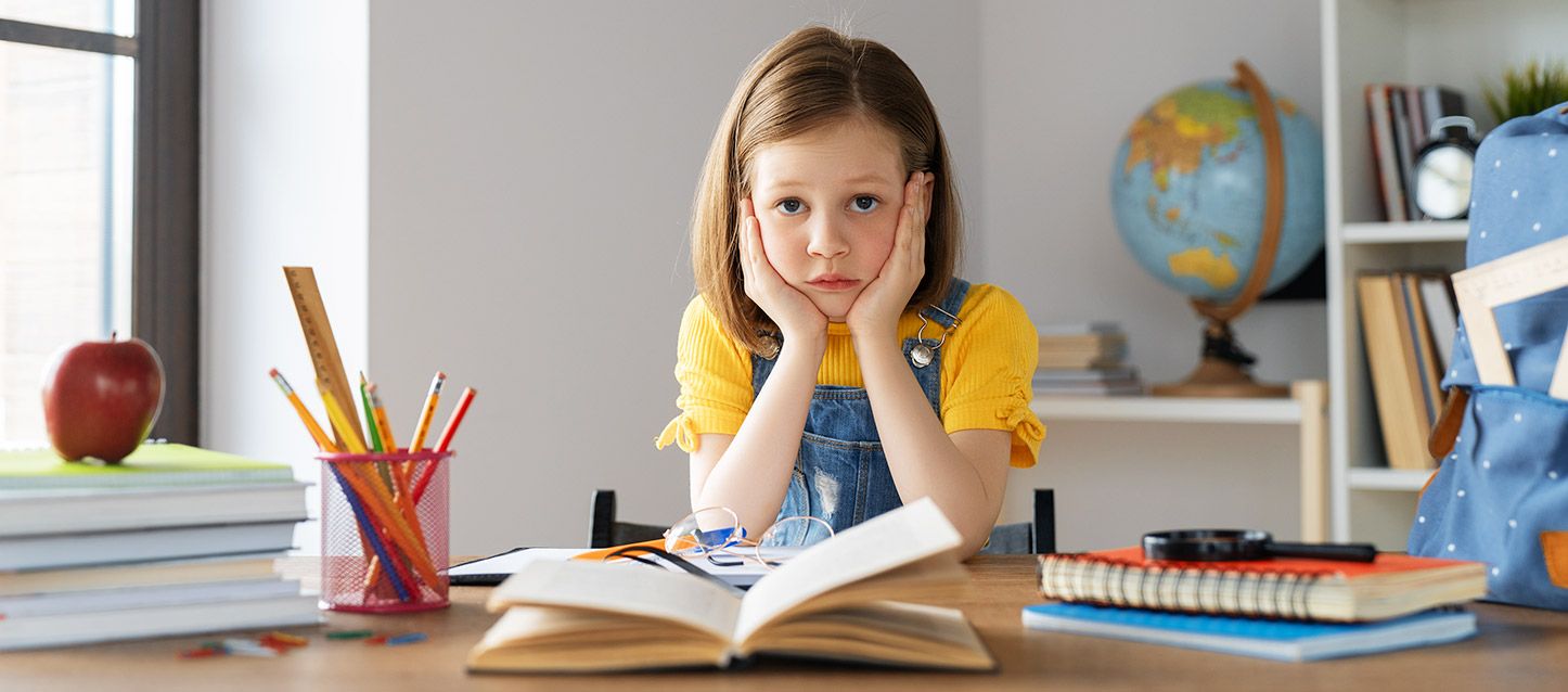 menina pequena com um macacão e uma camiseta amarela, a fazer os deveres, com as mãos na cara, e com um gesto de cansaço