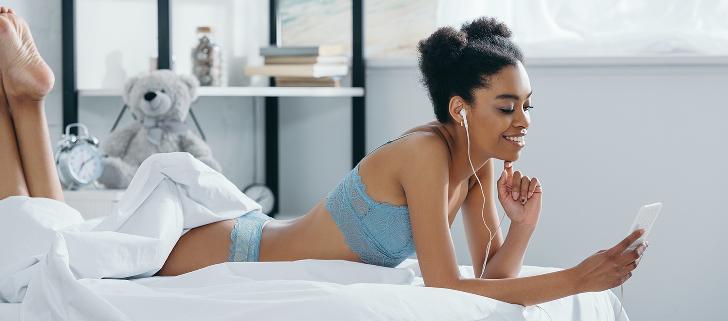 rapariga morena de cabelo afro apanhado com dois puxinhos, deitada de barriga para baixo na cama, a olhar para o telemóvel, com auriculares nos ouvidos