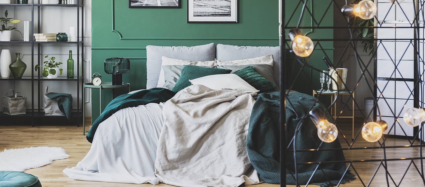 quarto com cama, edredão cinza, 6 travesseiros, 2 almofadas, parede verde, mesinha e estantes com objetos decorativos