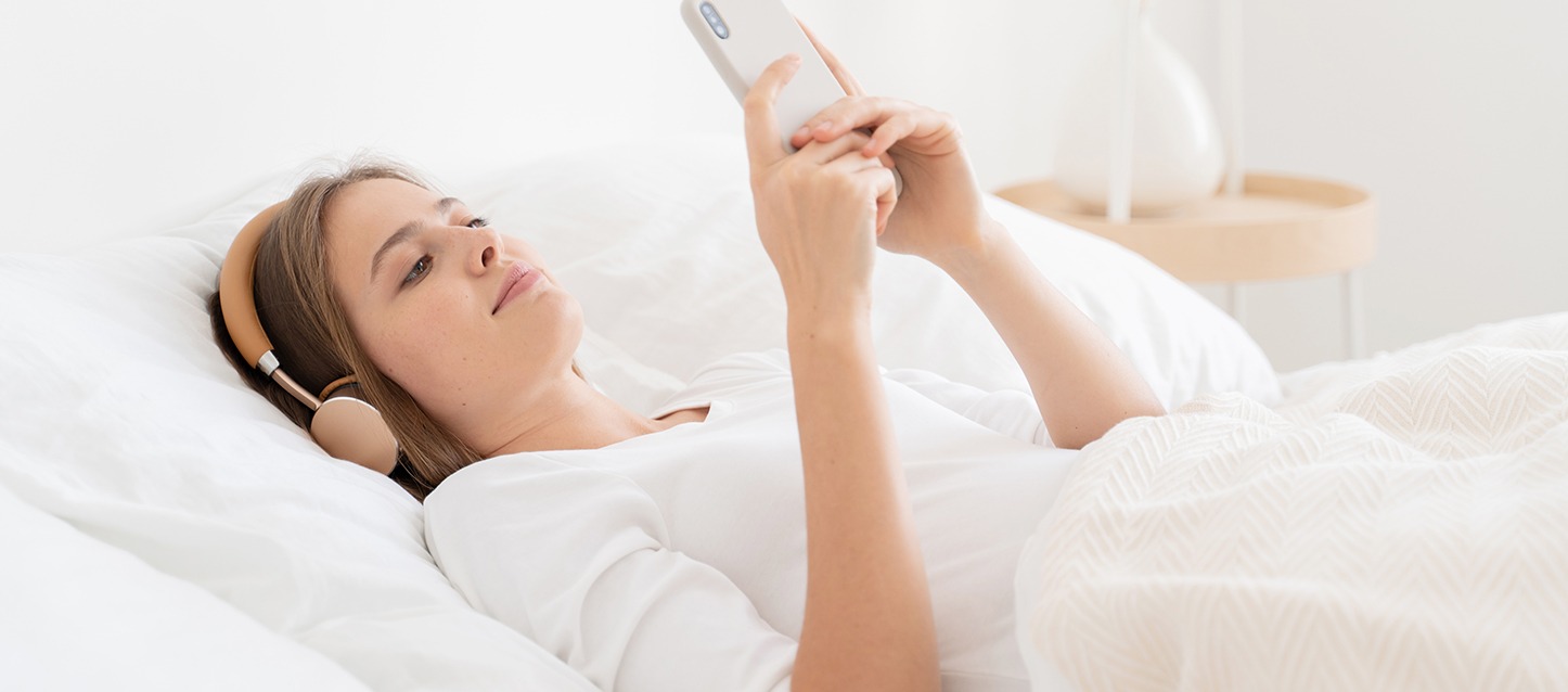 rapariga de cabelo castanho claro deitada na cama, com auriculares e a olhar para o telemóvel, a escutar ASMR