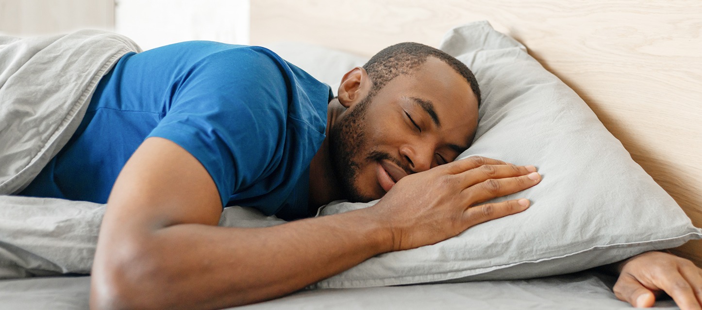Homem a dormir de barriga para baixo na cama e a sorrir.
