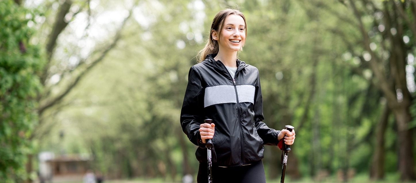 Mulher a correr marcha, com fato de treino preto e cinza, num parque com árvores.