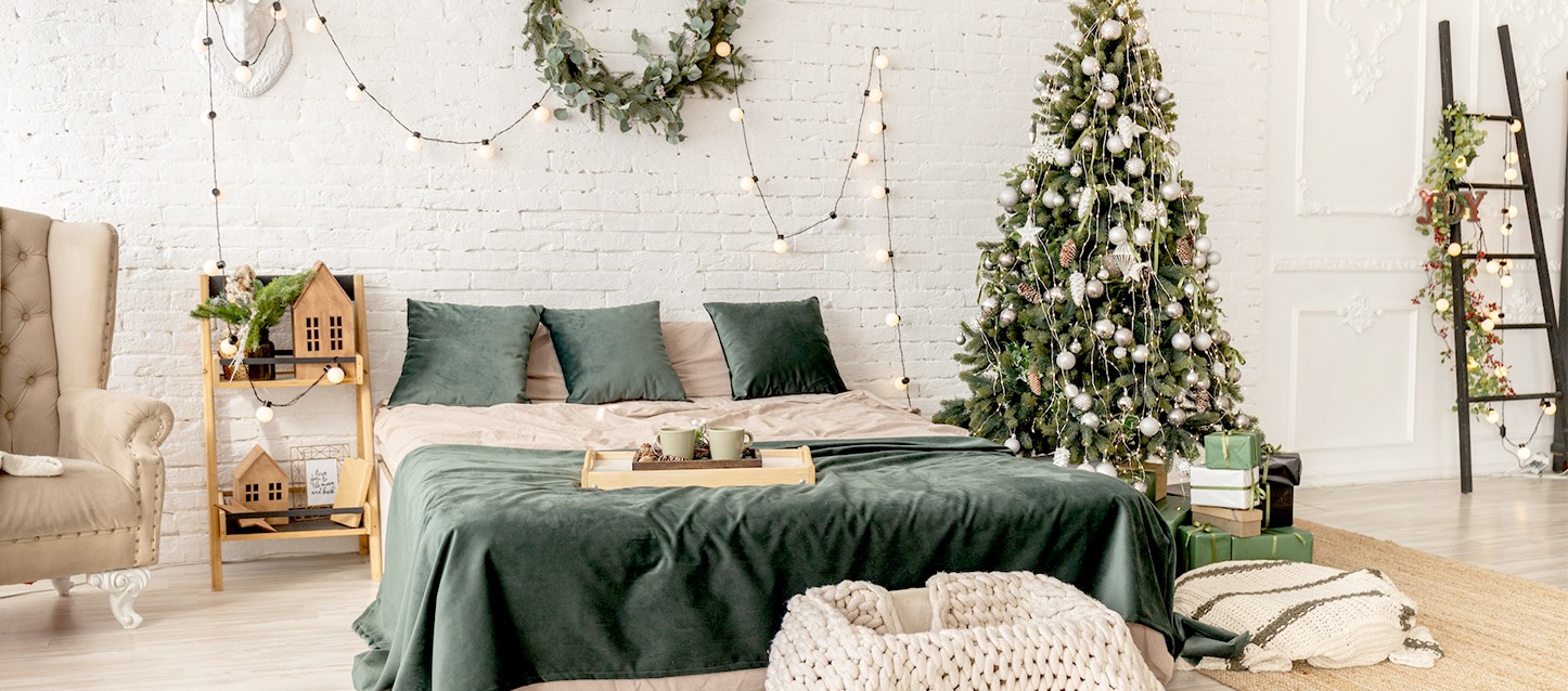 Quarto com paredes brancas, com uma cama com almofadas e travesseiros, árvore de Natal, mesinha de cabeceira e sofá.