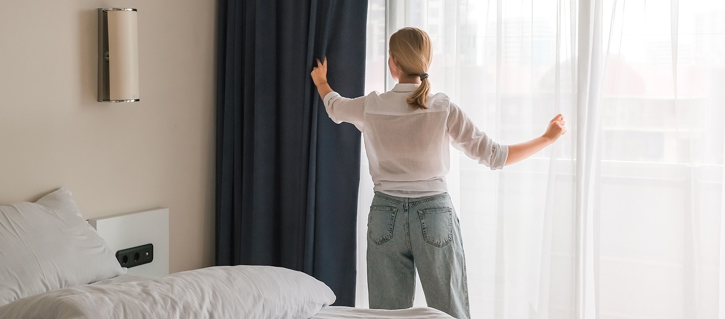 Mulher de cabelo loiro com camisa branca a segurar nas cortinas de um quarto.